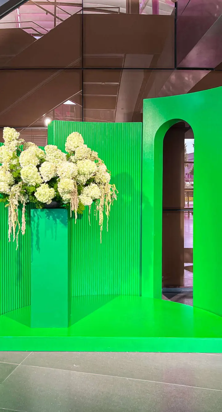 The image depicts a vibrant green display with large white floral arrangements, set against a backdrop of brown architectural elements.