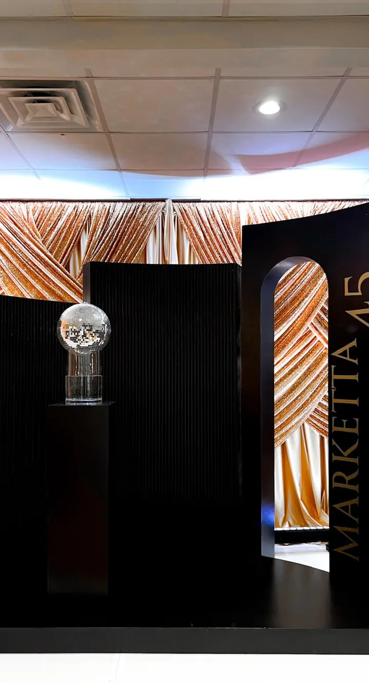 The image depicts a stage-like setup with a large black backdrop and a glass display case in the foreground, surrounded by a patterned fabric wall in warm tones.