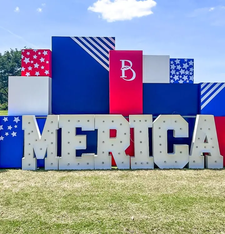 A large, colorful display featuring the word "MERICA" made up of various geometric shapes and patterns in red, white, and blue, set against a grassy field with trees in the background.