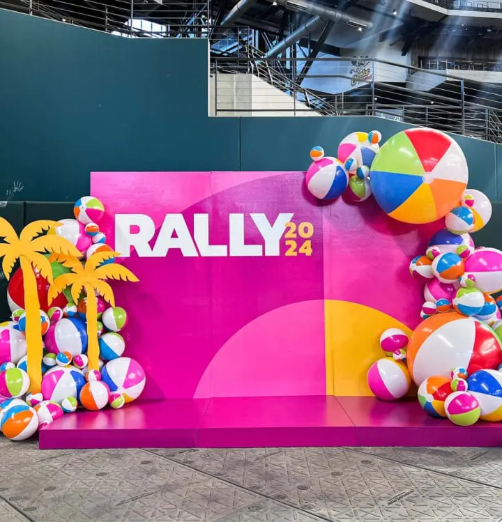 A vibrant and colorful display featuring a large "RALLY 2024" sign surrounded by inflatable beach balls, palm trees, and other playful elements against a backdrop of industrial structures.