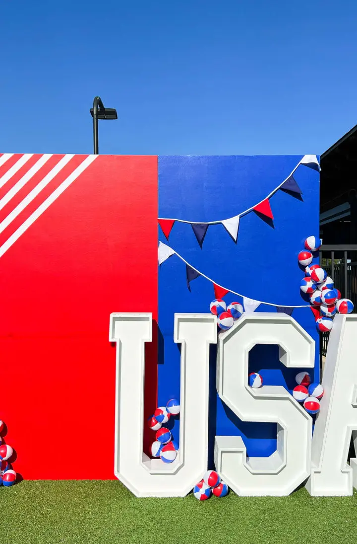 A large, colorful display featuring the letters "USA" surrounded by red, white, and blue balloons, set against a clear blue sky and a black metal fence in the background.