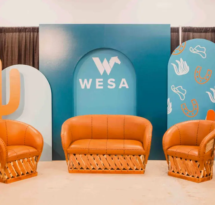 The image features a vibrant and modern setup with orange wicker chairs and a large blue backdrop displaying the "WESA" logo, surrounded by abstract shapes and patterns.