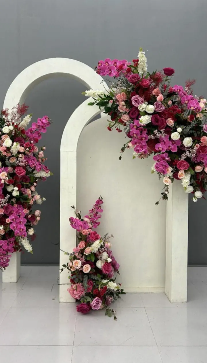 A white arched structure adorned with an abundance of vibrant pink and purple flowers, set against a plain gray background, creating a visually striking and elegant floral display.