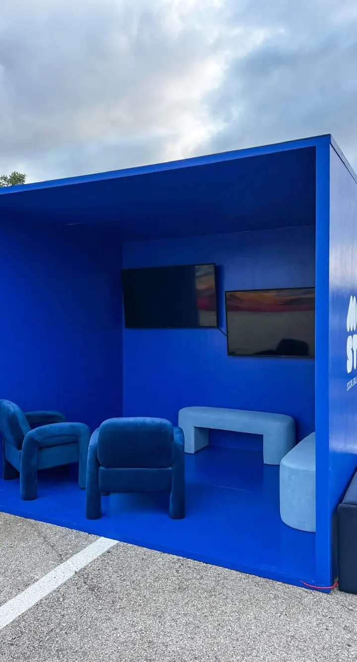 A bright blue modular structure with seating and a television screen, situated in an outdoor setting with a cloudy sky in the background.