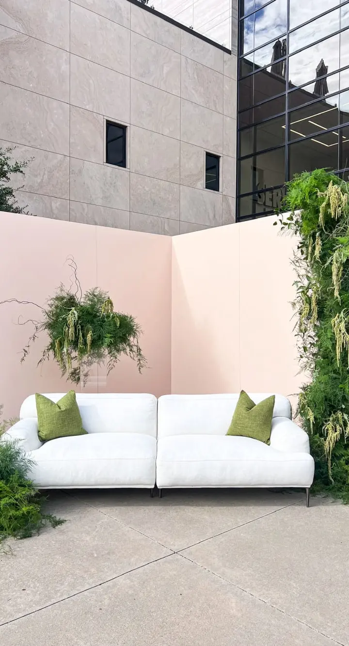 A modern, minimalist outdoor seating area with a white sofa surrounded by lush greenery and a concrete building in the background.