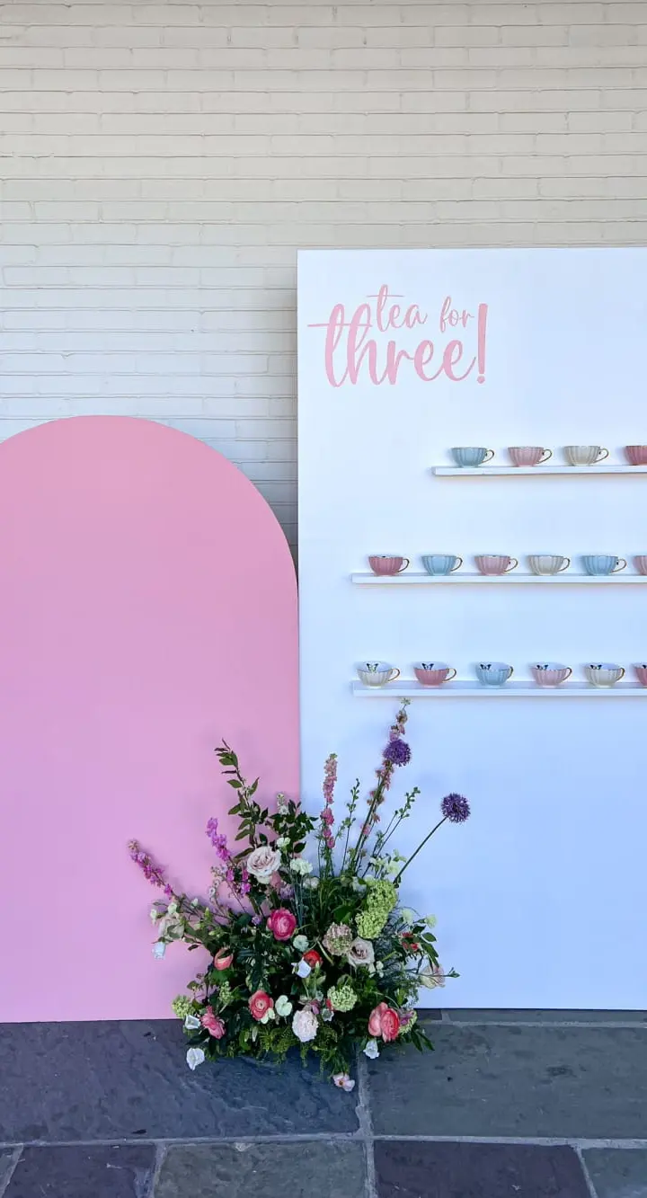 A large pink backdrop stands in the foreground, with a colorful floral arrangement placed in front of it. In the background, a white wall displays a sign that reads "Tea for three!" and features a display of various teacups.