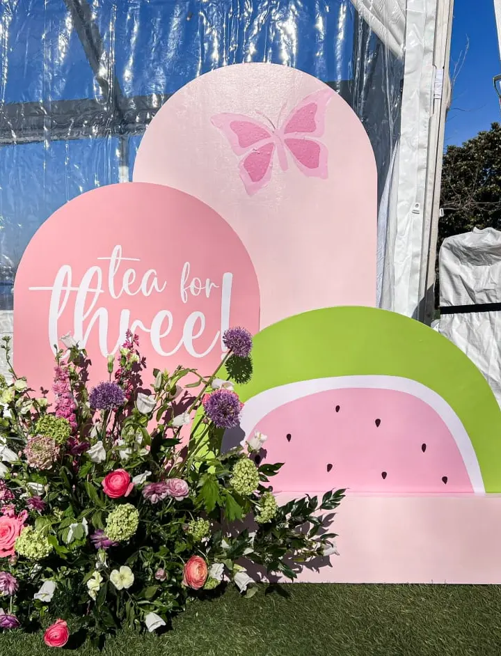The image shows a display of large pink and green shapes resembling watermelons and butterflies, with a bouquet of colorful flowers in the foreground, set against a backdrop of a building and blue sky.