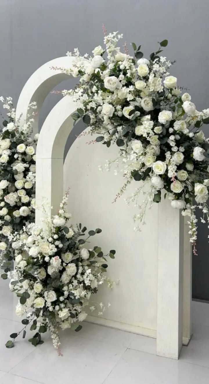 A white, arched floral arrangement filled with delicate white flowers and greenery sits on a plain white surface against a gray background.